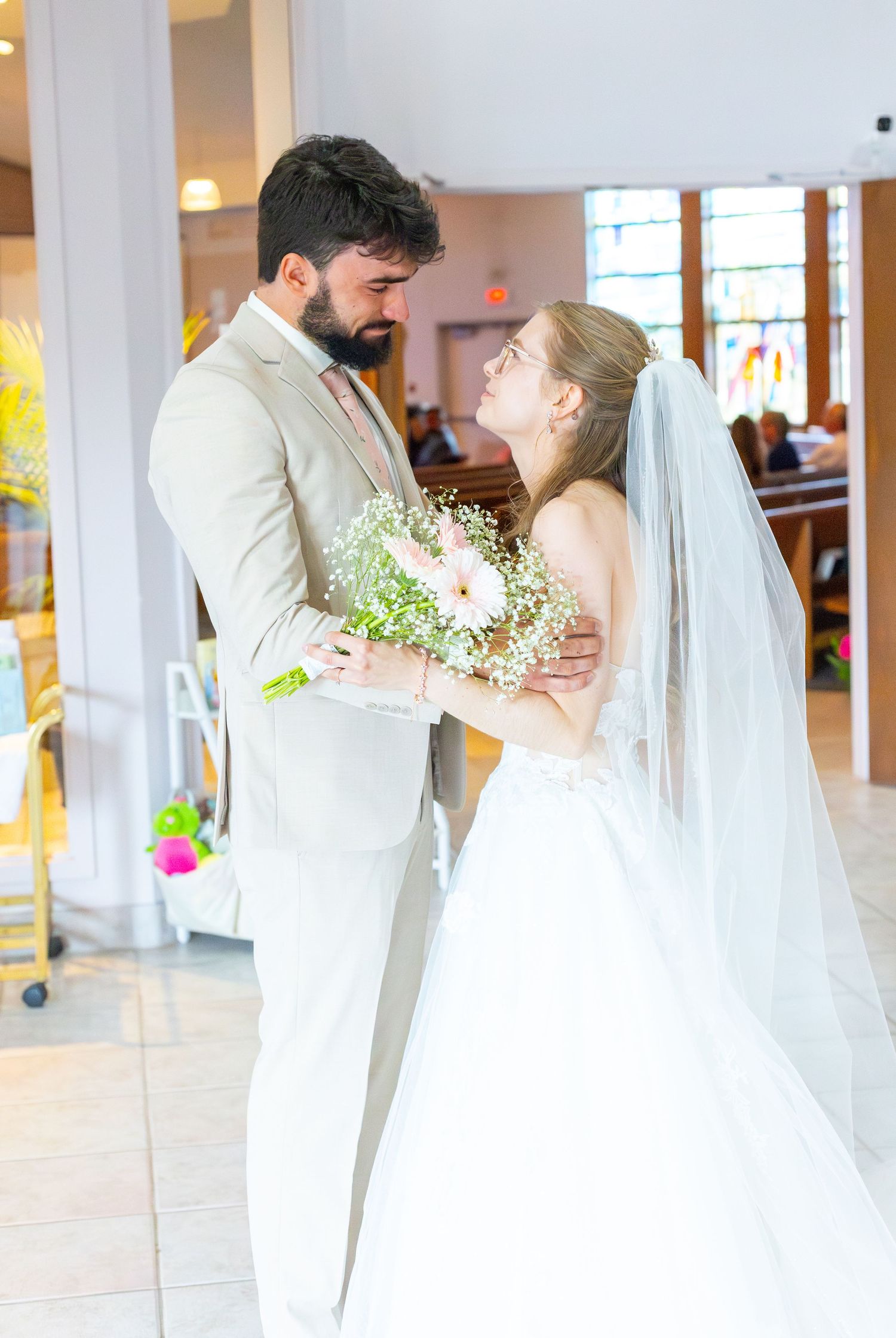 Groom in beige suit embracing bride in white gown holding bouquet inside a bright church.