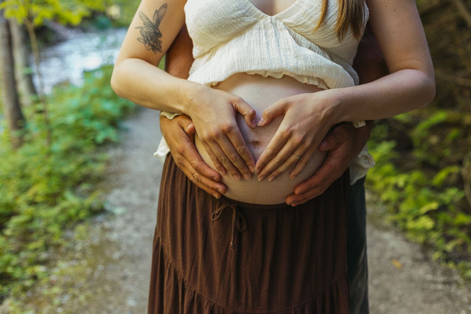 Close-up of a pregnant woman with tattoos forming a heart shape with her partner's hands on her belly.