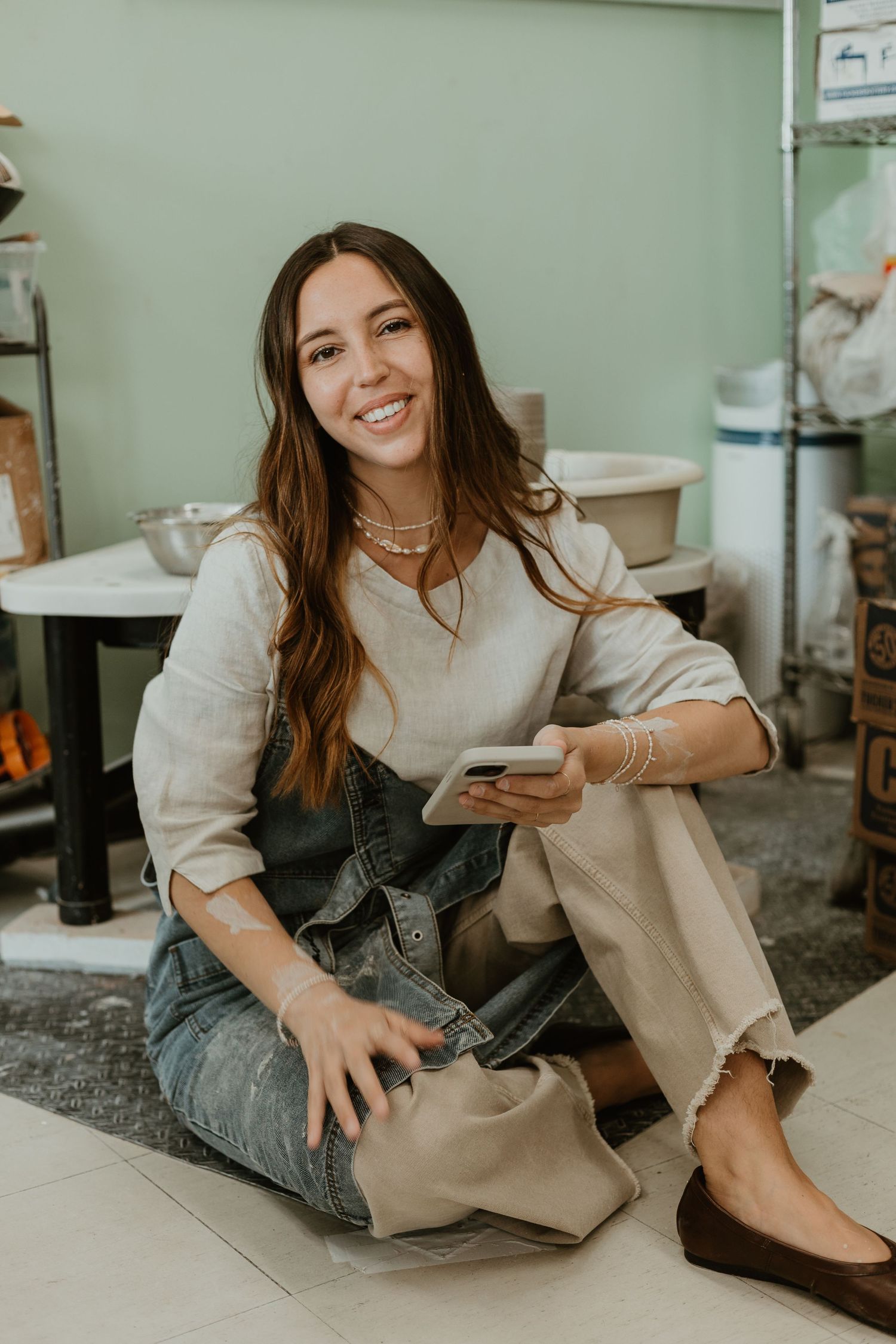 Smiling woman sitting on floor in pottery studio, holding smartphone, wearing linen top, denim apron, and tan pants.