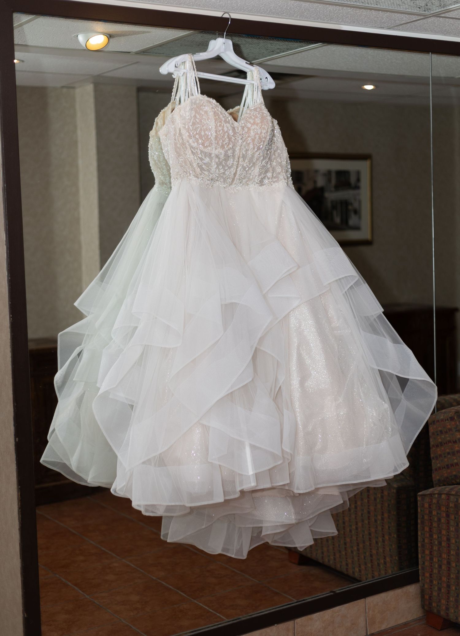 Elegant layered ruffled white wedding gown with lace bodice hanging on a mirror in a hotel room.
