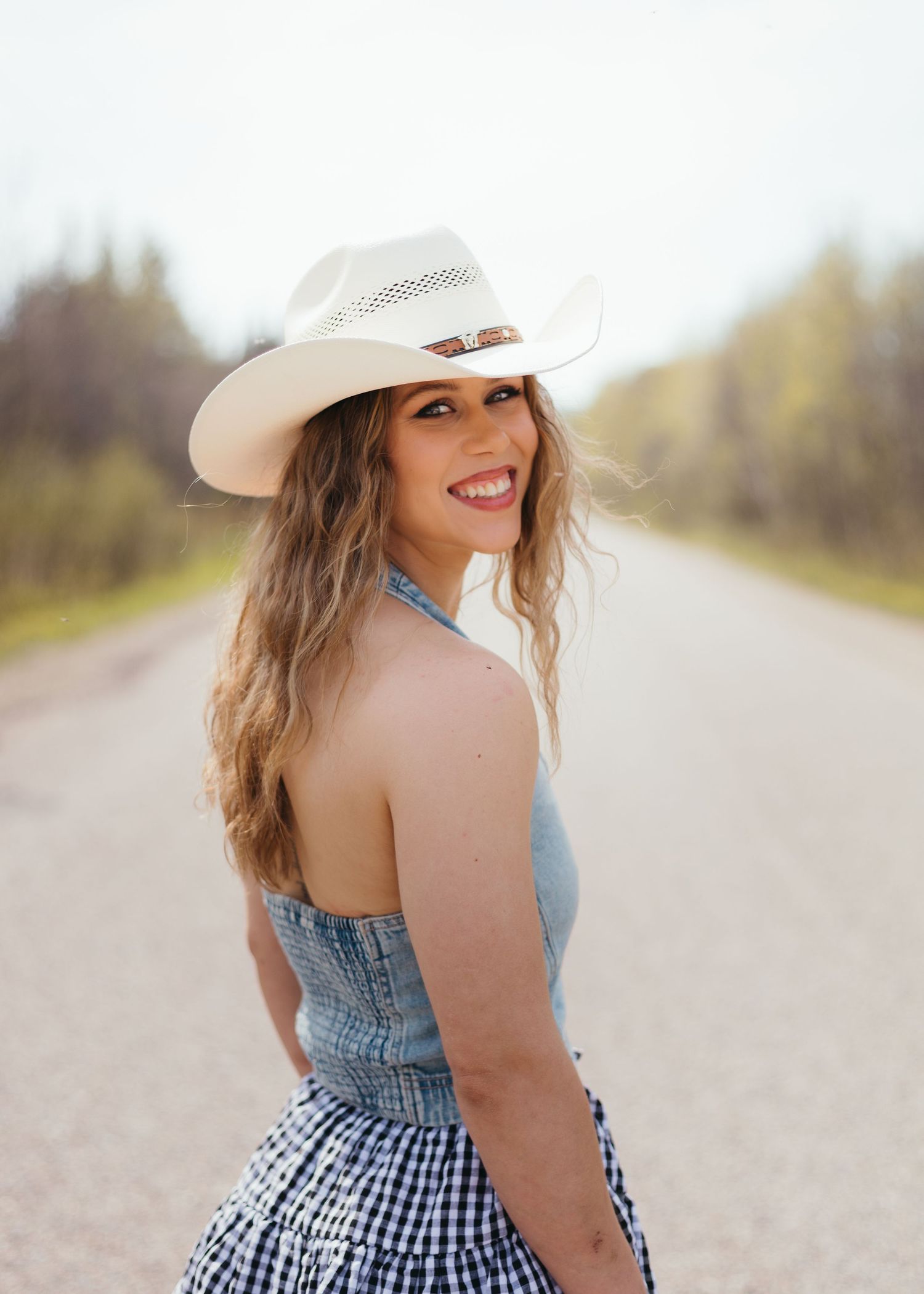 Smiling woman in white cowboy hat and denim gingham dress poses on a rural road with blurred trees in background.