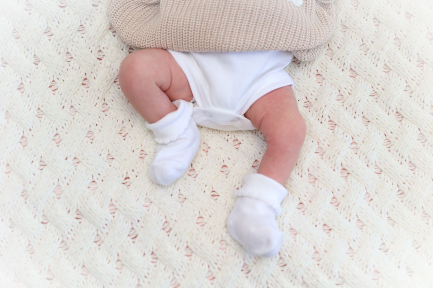 Close-up of a newborn baby's legs and feet in white socks lying on a cream quilted blanket.