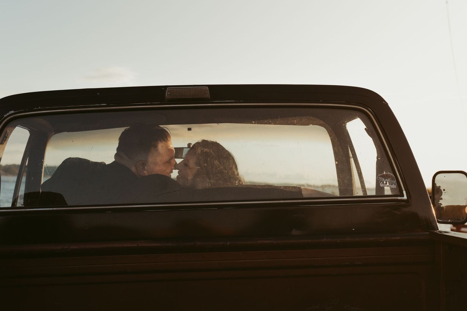 Romantic couple sharing a kiss inside a classic vintage truck cab at sunset by the water.