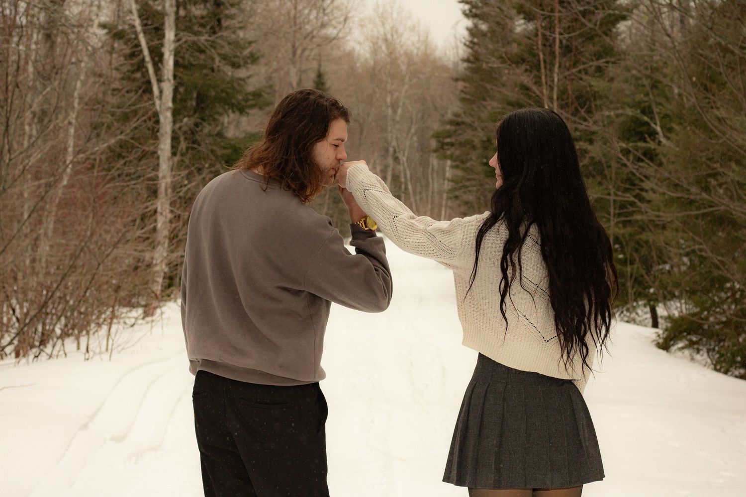 A couple stands in a snowy forest, man kissing woman's hand in a romantic winter gesture.
