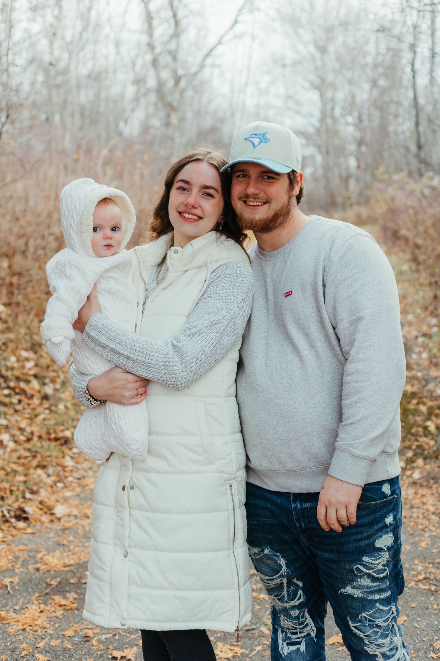 Young family with baby poses on autumn path in white puffer coat and gray sweatshirt.