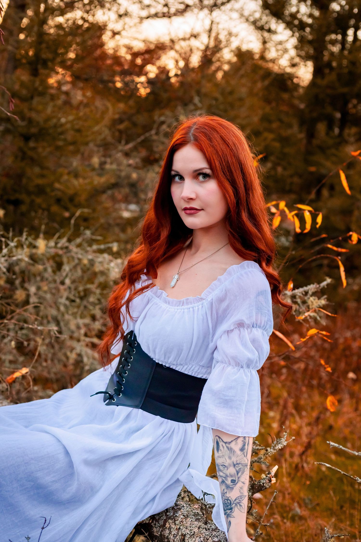 A woman with long red hair wearing a white dress and black corset sits outdoors in an autumn forest setting.
