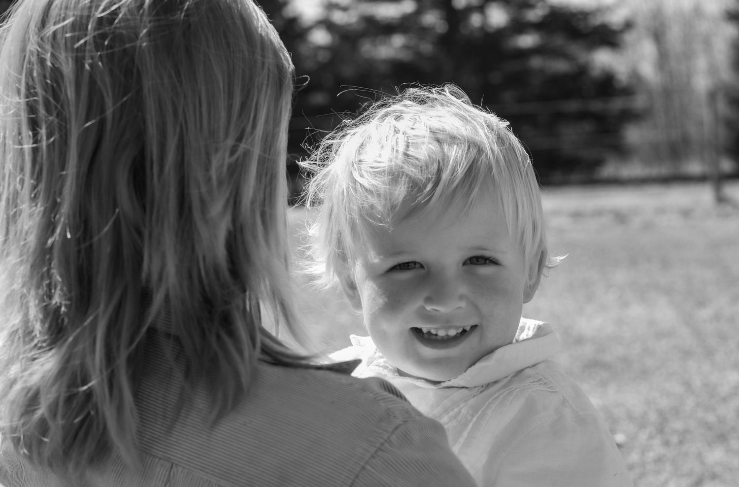 Black and white photo of a smiling toddler being held by a woman outdoors in a park.