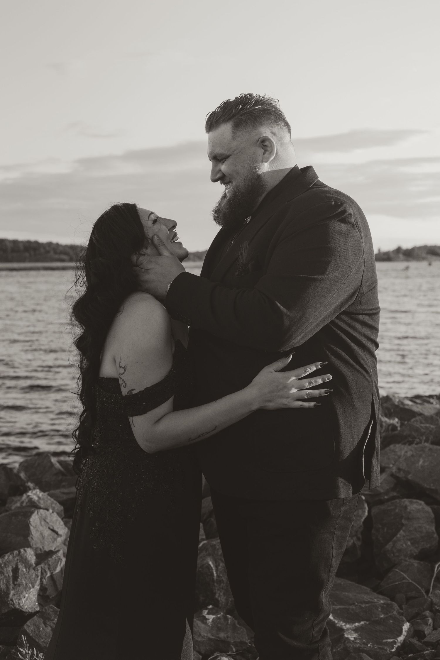 Black and white romantic photo of couple embracing by rocky waterfront at sunset after wedding.