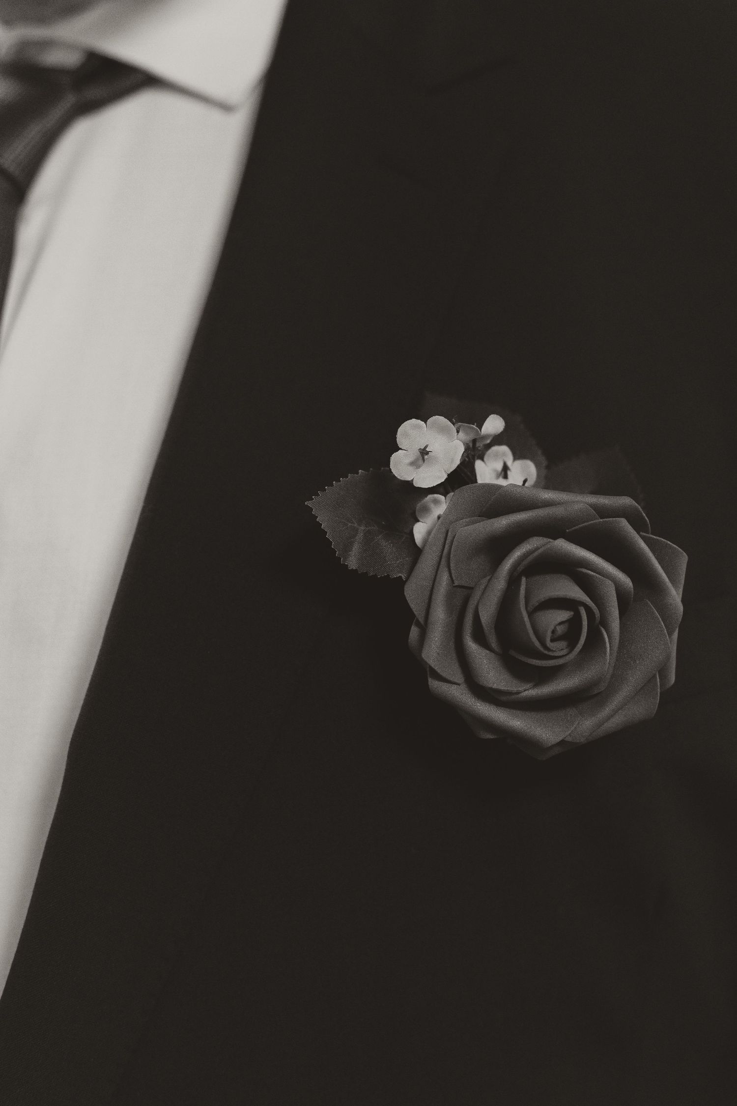 Black and white close-up of a groom's boutonniere with a rose and small white flowers on suit lapel.