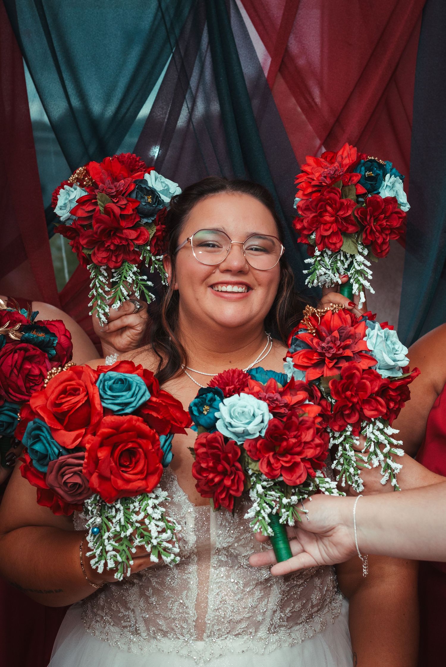 Smiling bride in glasses holds red and teal bouquets surrounded by bridesmaids at colorful backdrop.