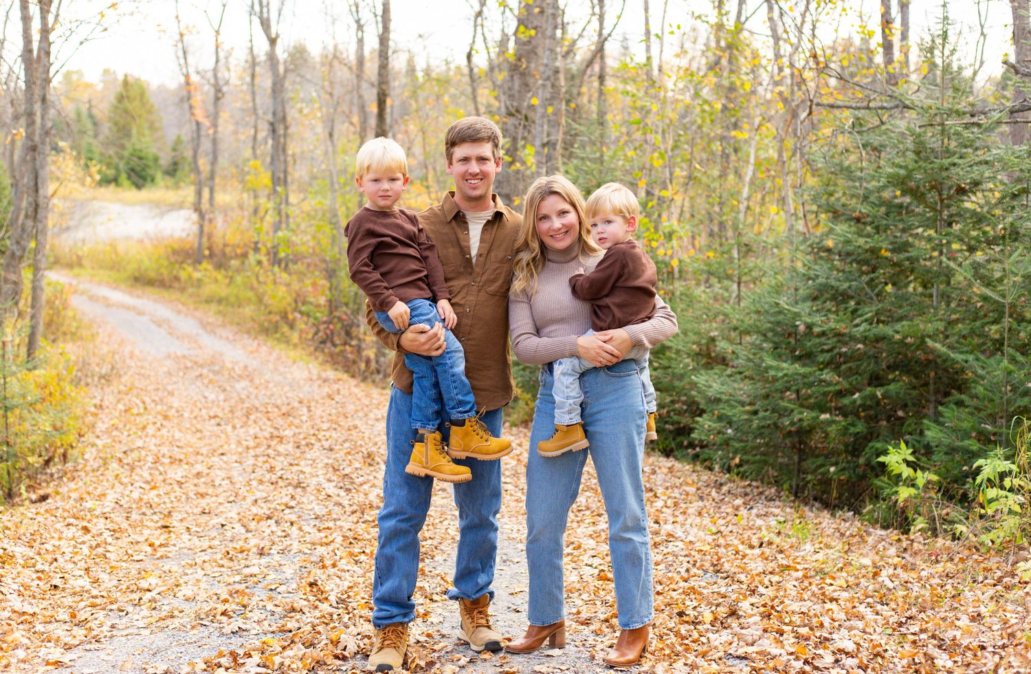 Young couple holds two toddlers on leaf-covered autumn trail lined with pine trees.