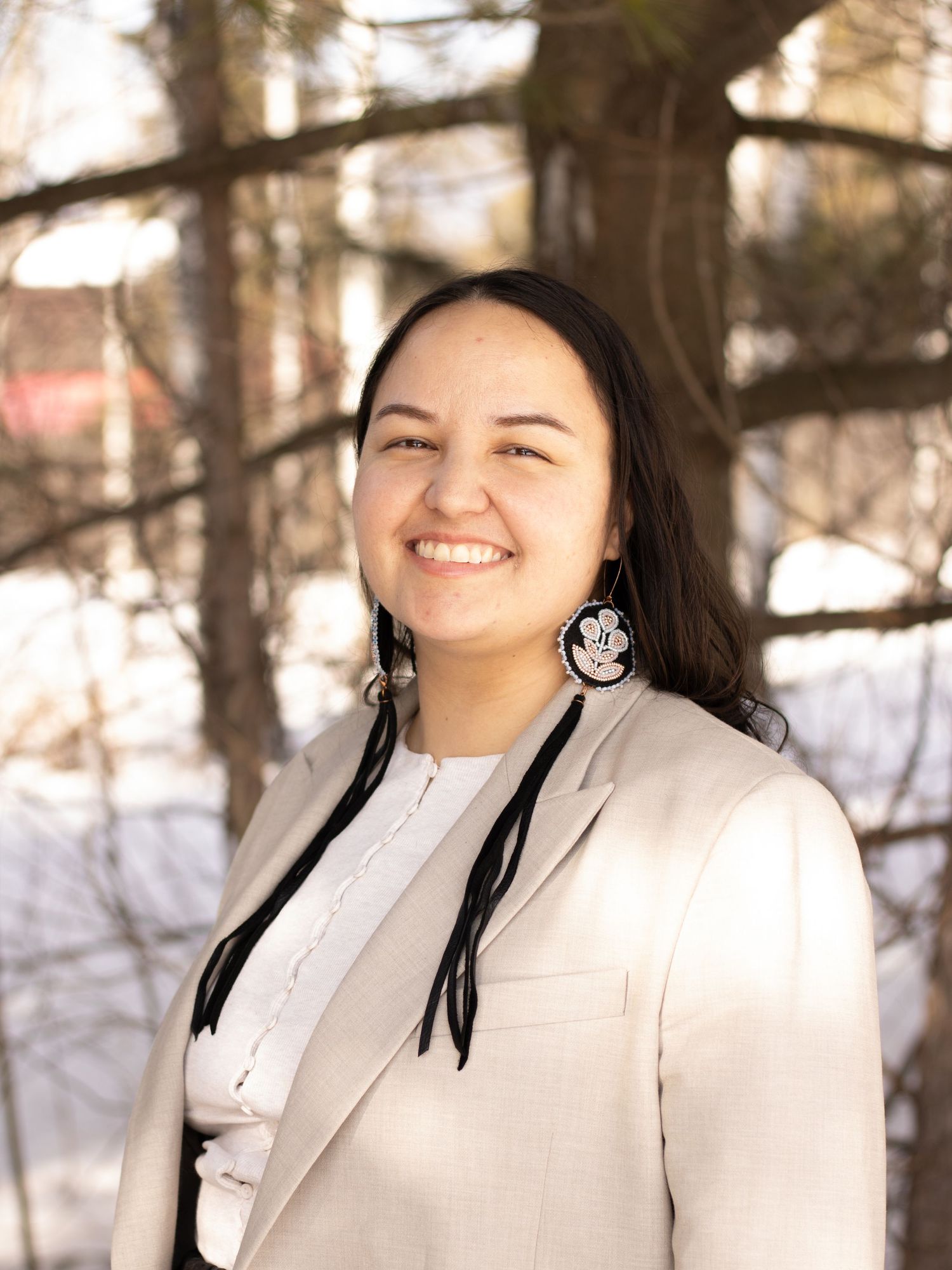 A young woman with dark hair in beaded earrings and a beige blazer smiles in a snowy outdoor setting.