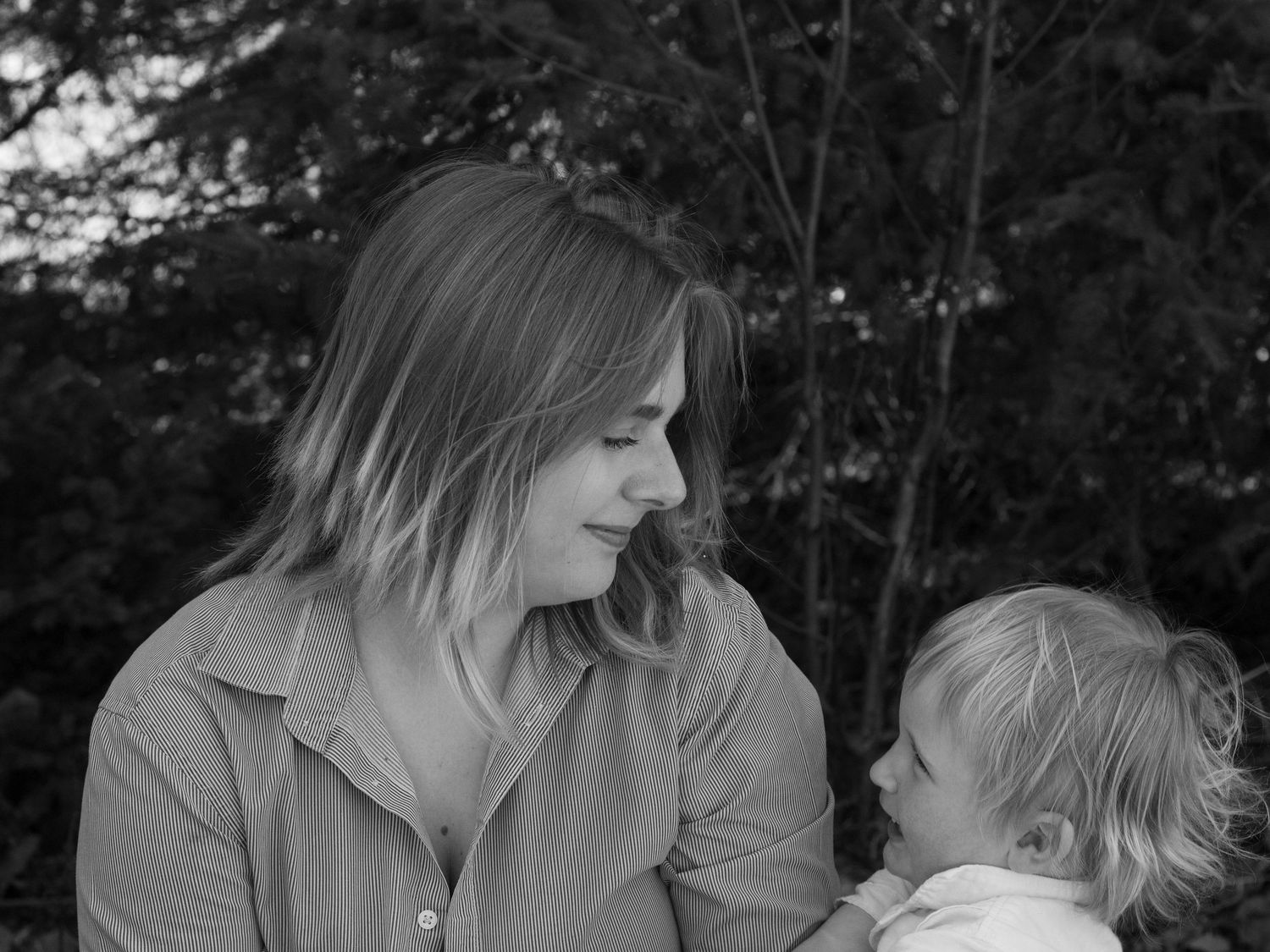 Black and white portrait of a mother smiling at her young toddler child outdoors among trees.