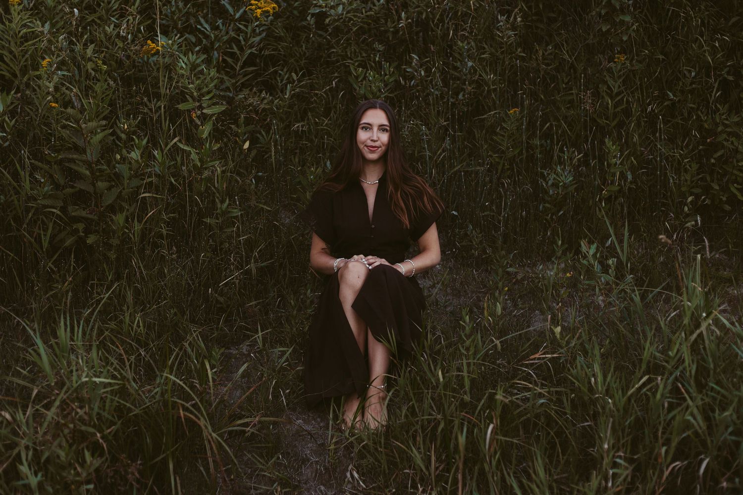 Brunette woman in black dress sitting peacefully among lush green wildflowers and tall grass.