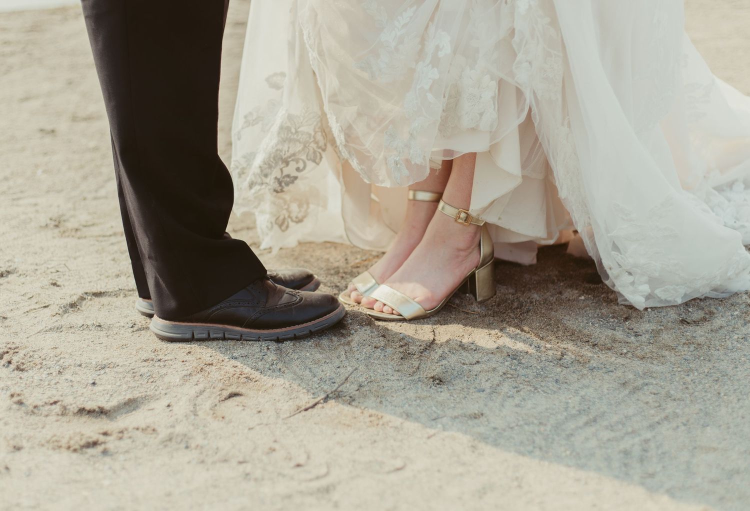 Close-up of bride in lace gown and groom in black pants standing on sandy ground.