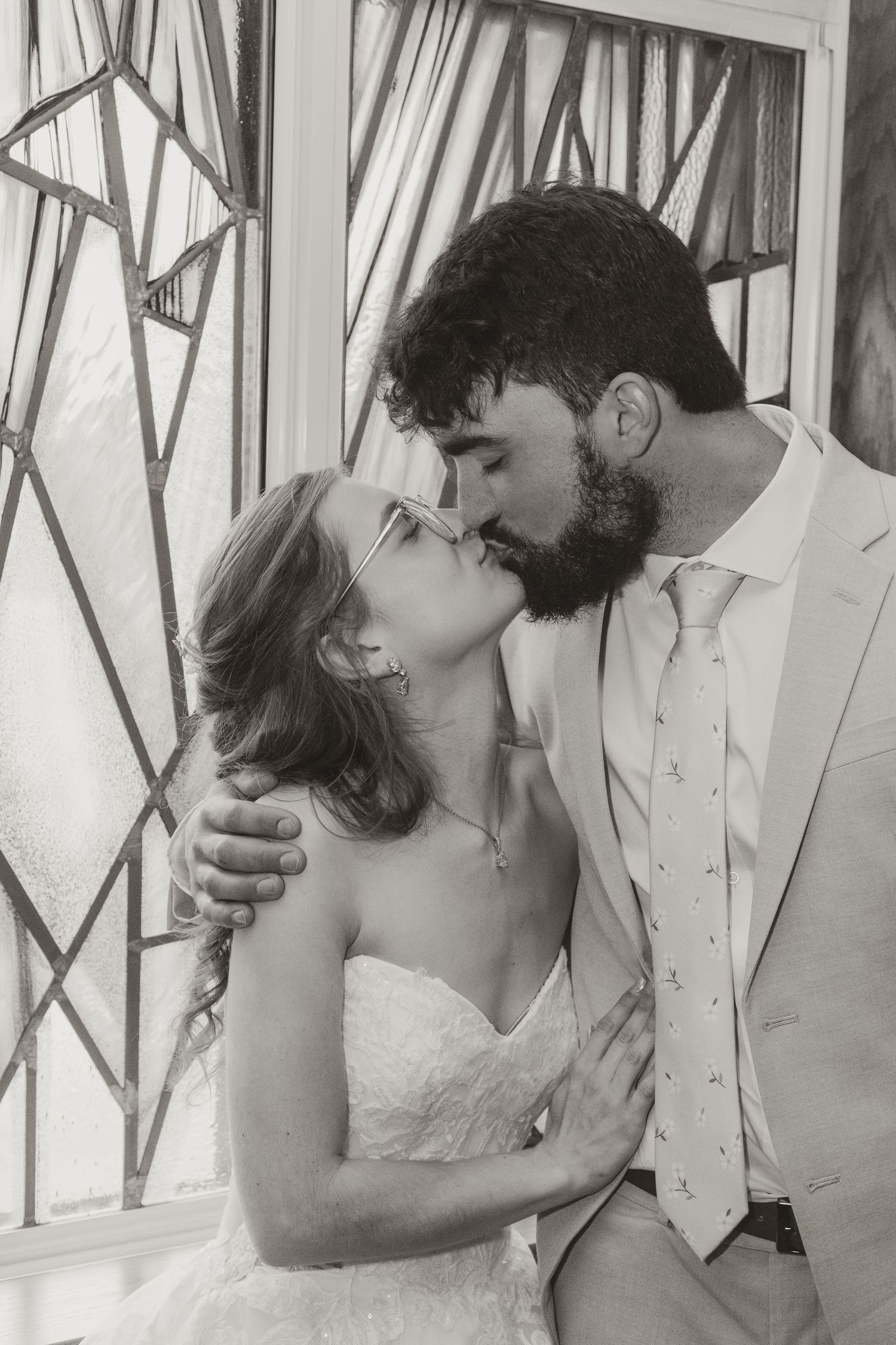 Black and white photo of bride and groom sharing a kiss near stained glass windows on their wedding day.