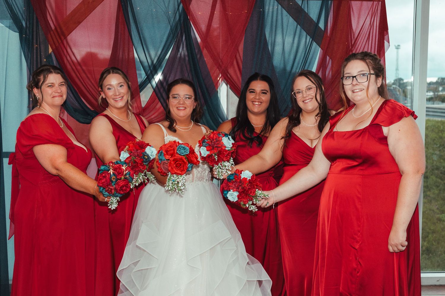 Bride in white gown with five bridesmaids in red dresses holding bouquets of red and blue flowers at wedding.