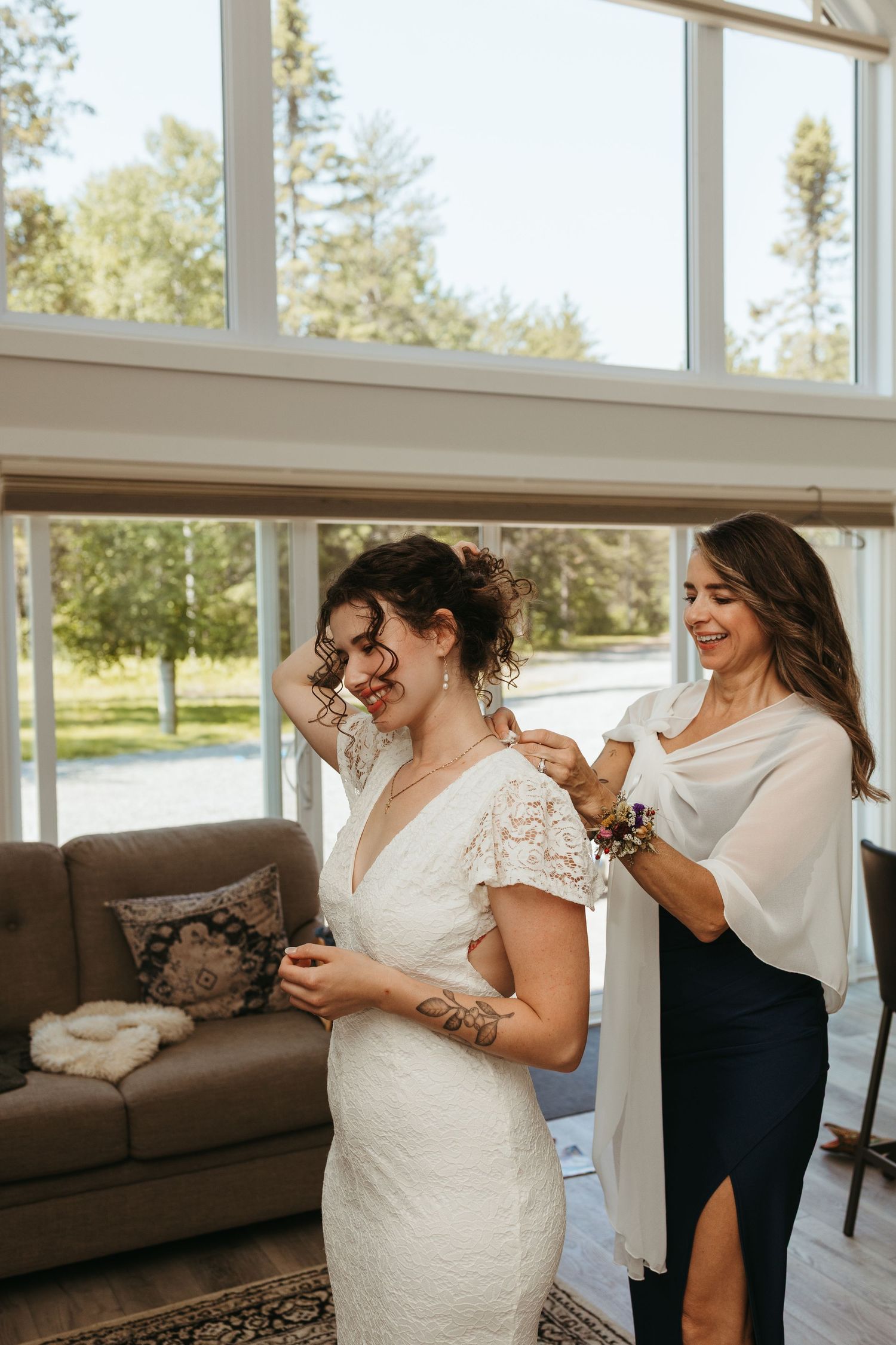 A bride in a white lace gown gets help from a smiling bridesmaid in a navy dress inside a bright, modern venue.