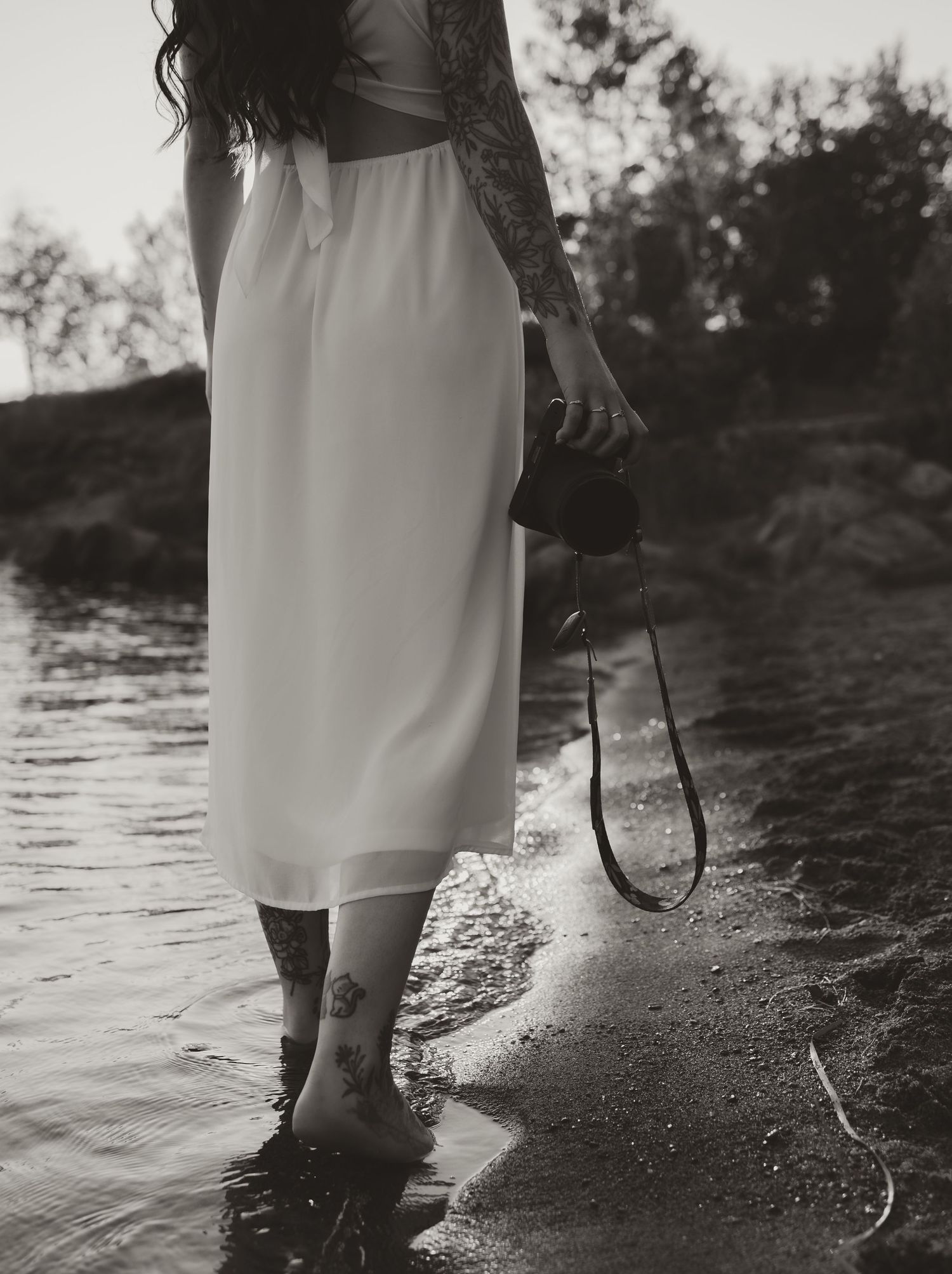 Black and white photo of tattooed woman in white dress holding camera while wading in shallow water near shore.