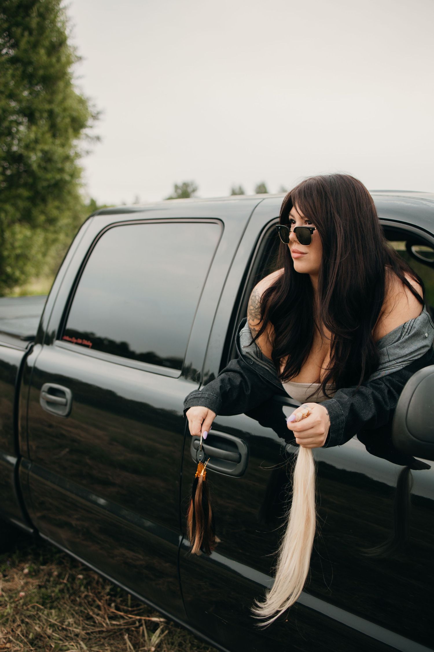 Dark-haired woman in sunglasses leans out of black pickup truck window holding long blonde hair extensions.