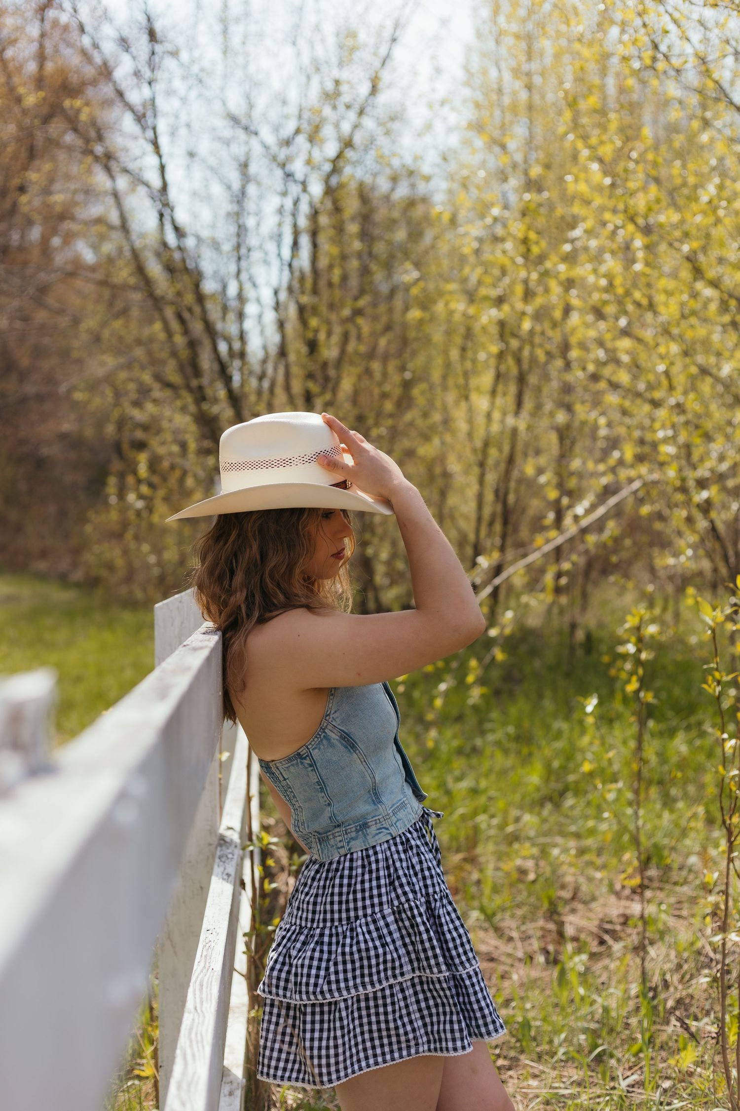 Young woman in denim vest, plaid skirt, and straw hat leaning on white fence surrounded by spring trees.