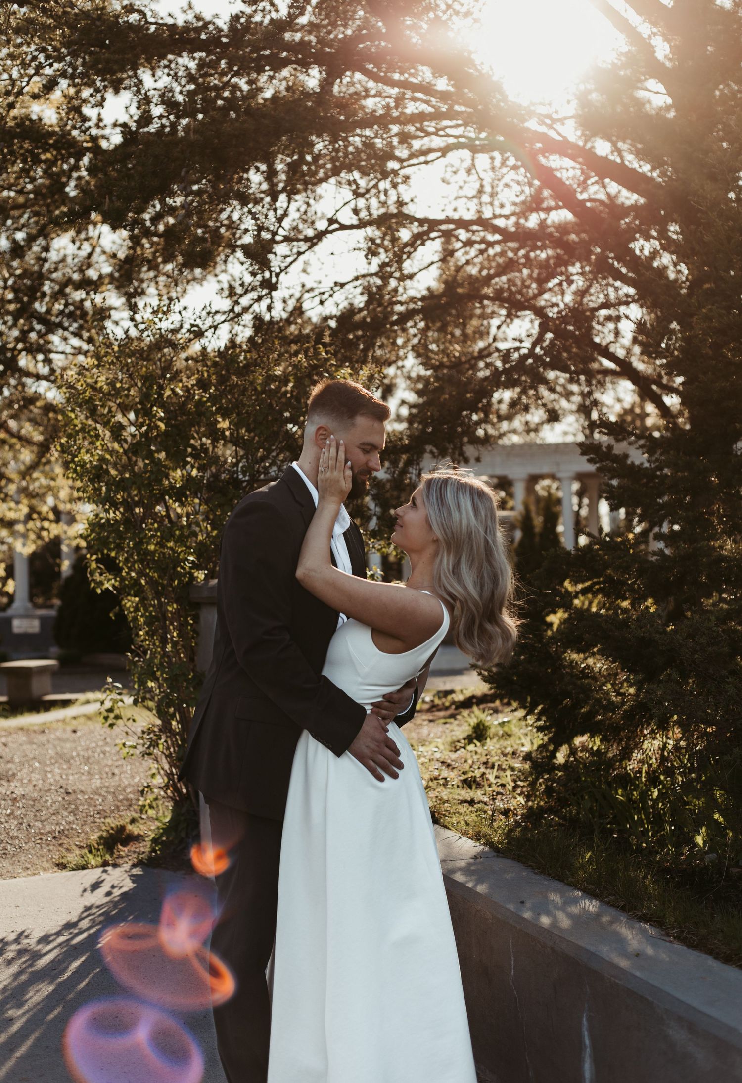 A couple shares a romantic moment in golden sunset light, the bride in white gown and groom in dark suit embracing outdoors.