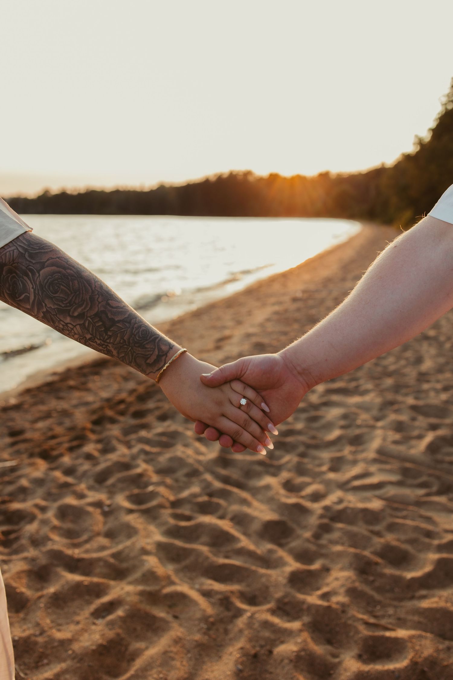 Couple holding hands on sandy beach at sunset, one with tattooed arm and other wearing diamond engagement ring.