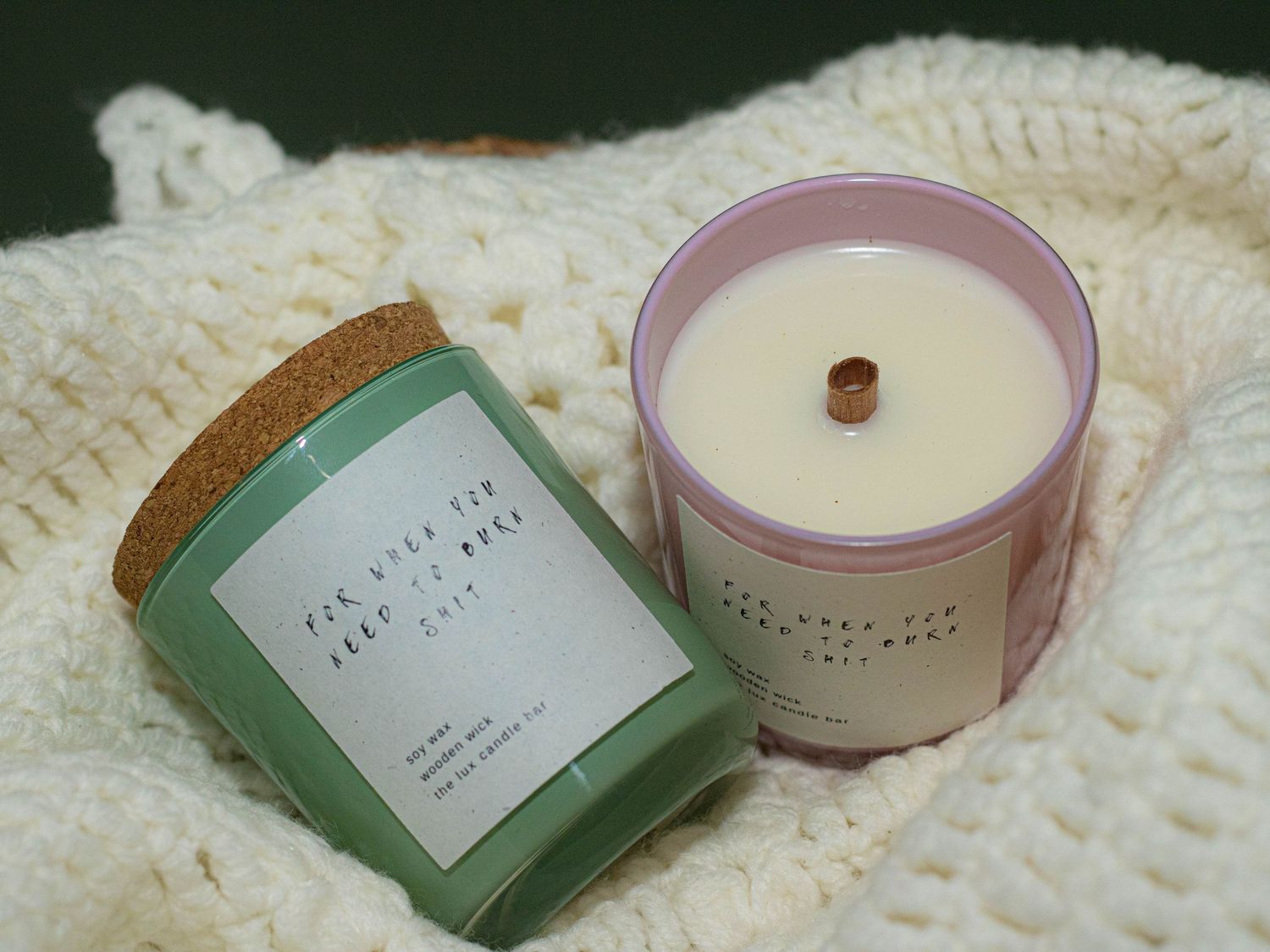 Two handmade candles with cork and wood wicks resting on a chunky knit white blanket.