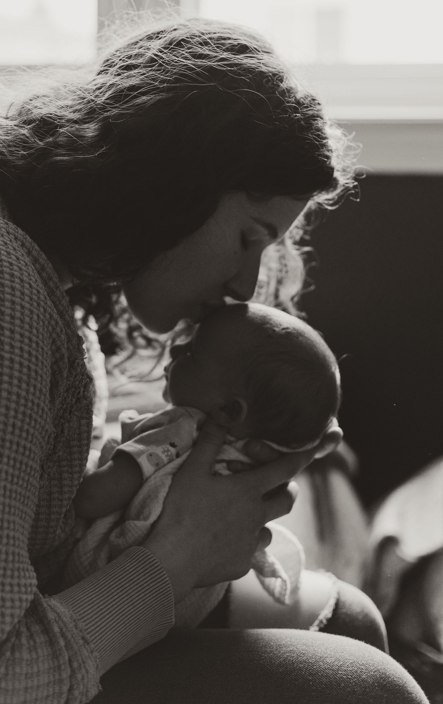 Black and white photo of mother gently kissing her newborn baby in soft window light.