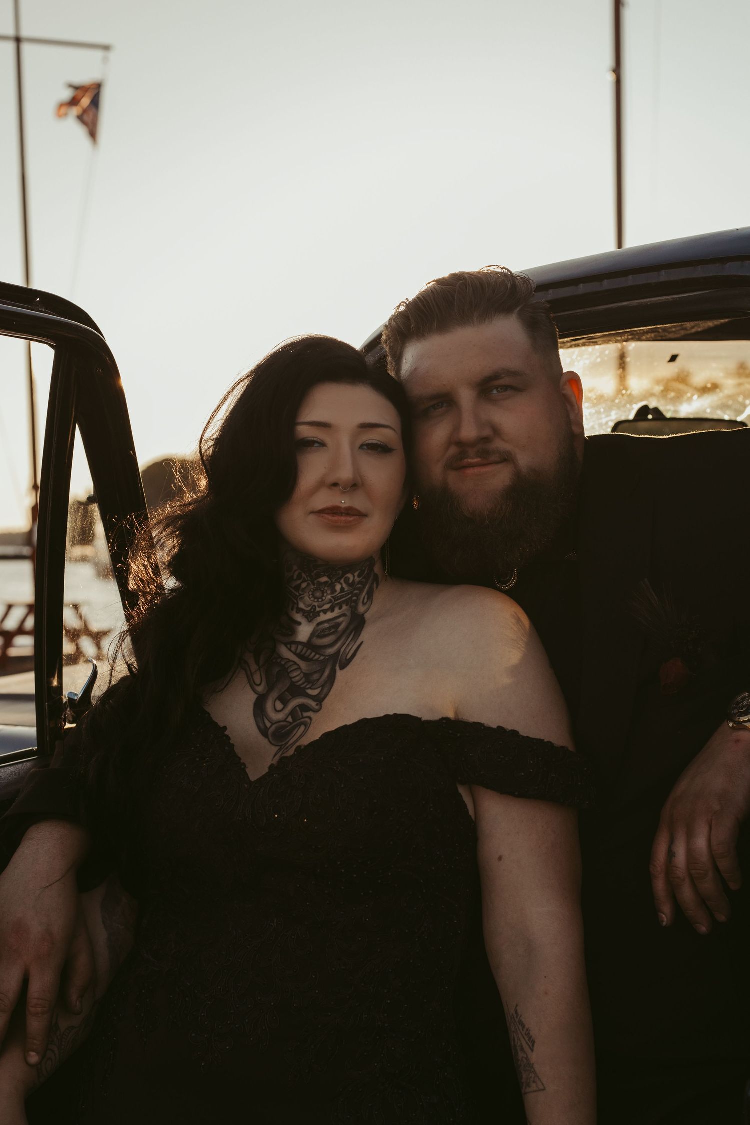 Couple in black outfits posing in vintage car, woman with neck tattoos leaning on bearded man at golden hour.