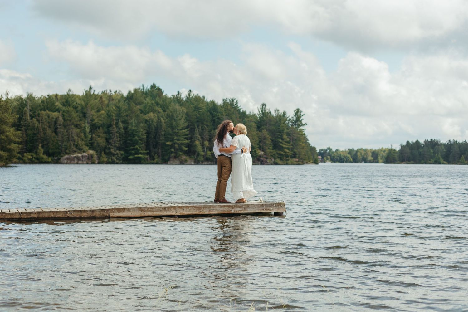 Couple sharing a romantic kiss on a wooden dock surrounded by a serene lake and lush forest backdrop.