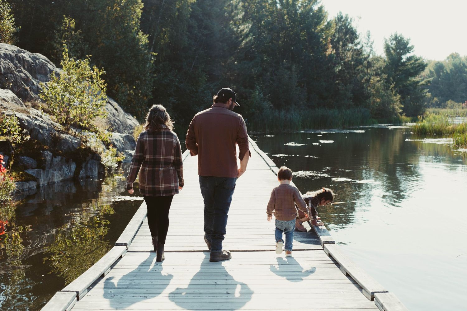 Family with toddler walks along wooden dock beside calm reflective lake.