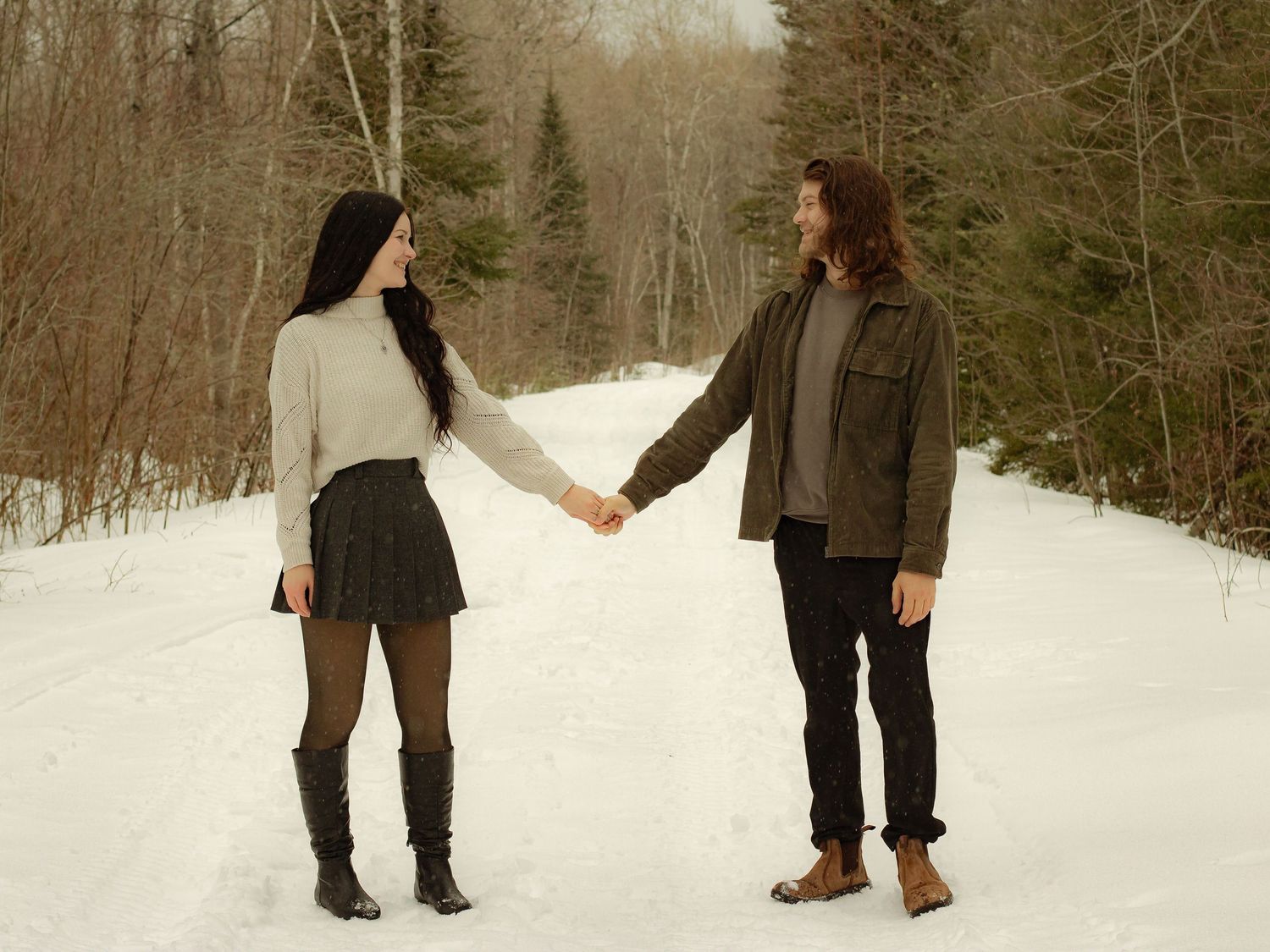 Couple holding hands on a snowy winter road surrounded by snow-covered pine trees.