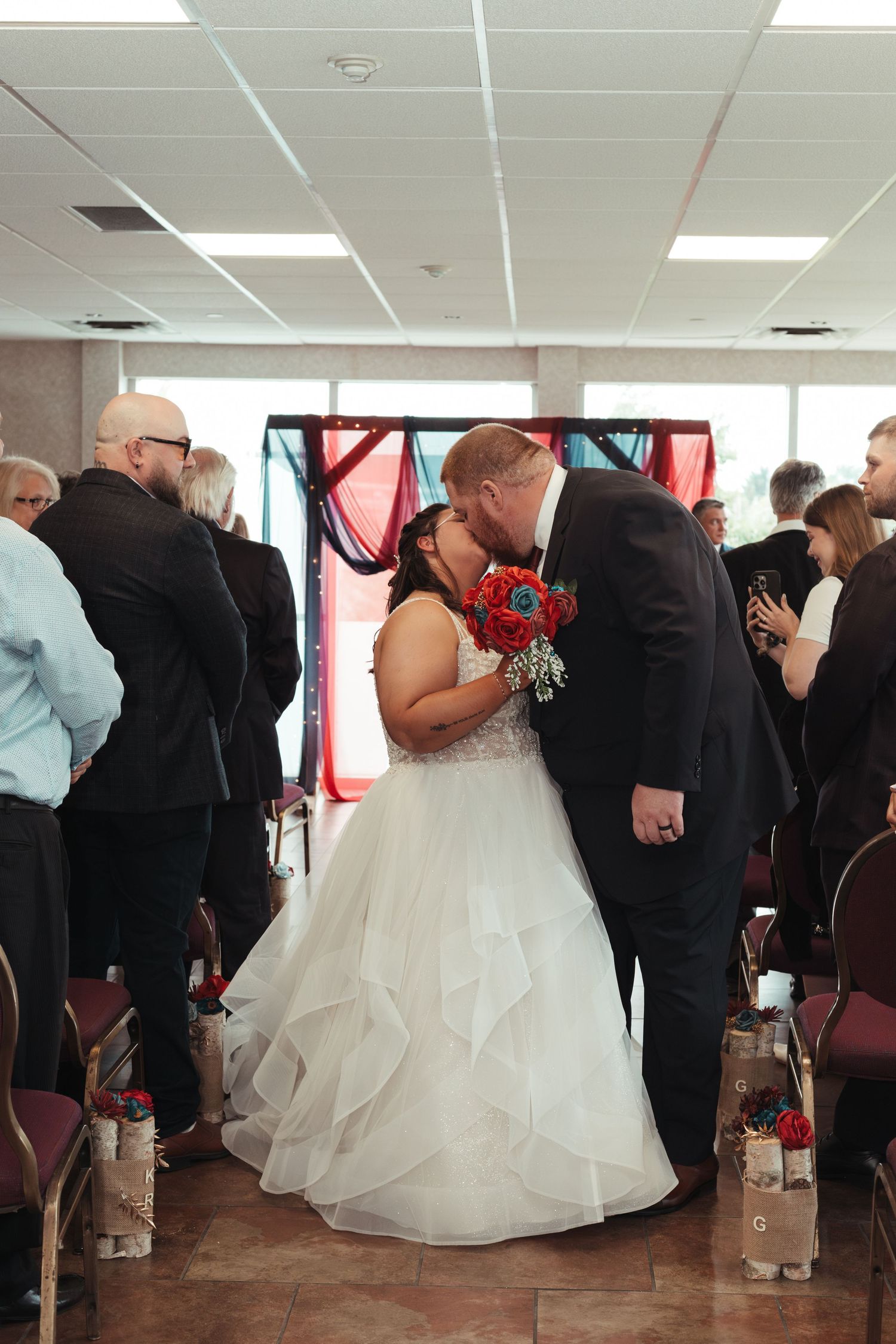 Bride and groom share first kiss at the altar surrounded by guests during indoor wedding ceremony.
