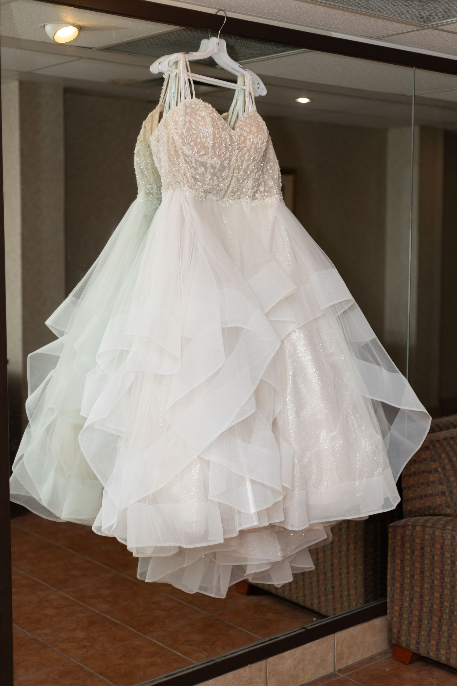 Elegant white layered tulle and lace wedding gown hanging on a mirror in a hotel room.