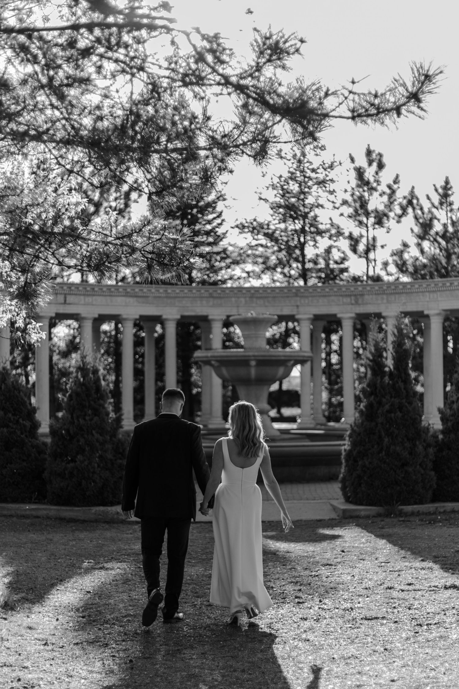 Black and white photo of bride and groom holding hands walking toward elegant fountain colonnade in lush garden.