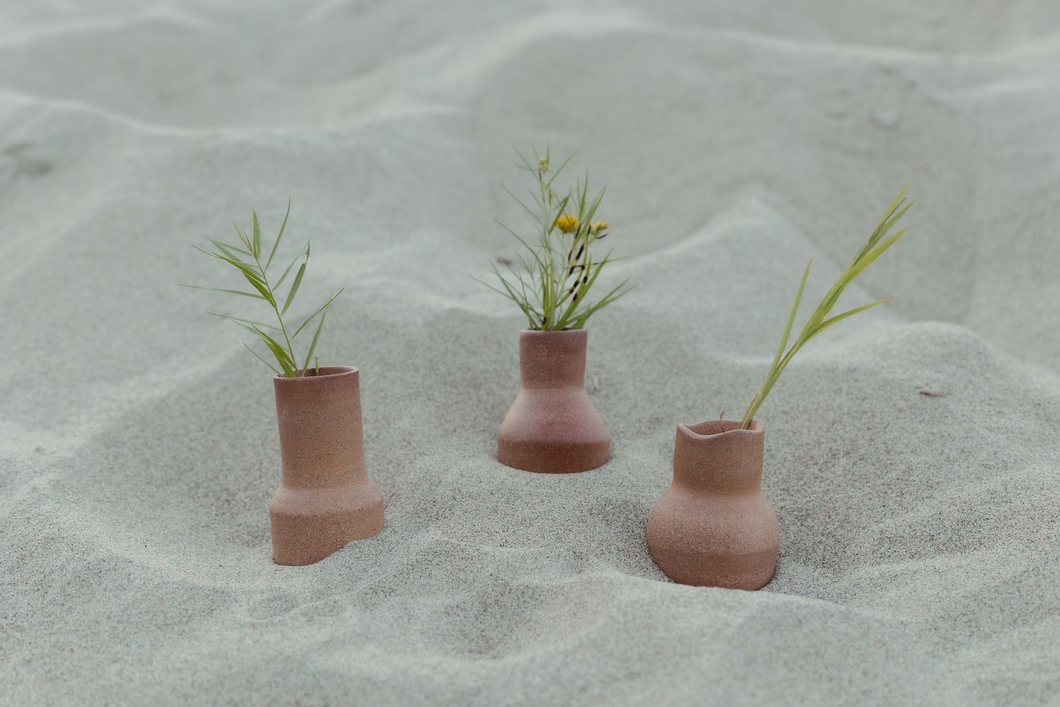 Three small terracotta vases with wildflowers arranged in white sand at beach setting.