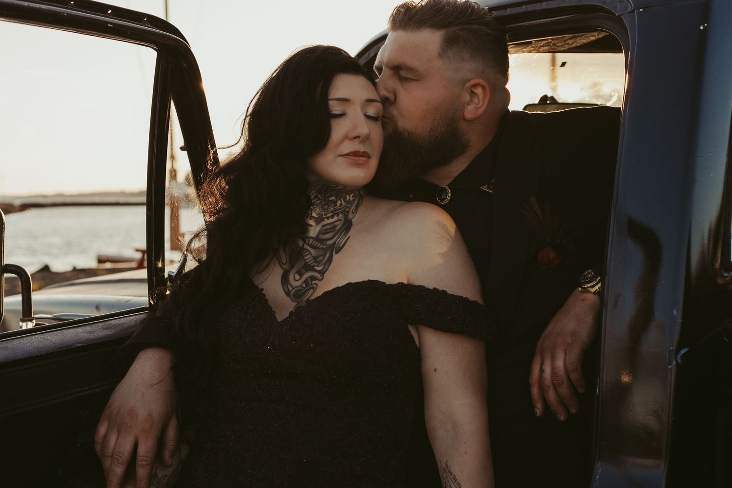 Romantic couple embracing in vintage truck, woman with neck tattoo in black dress, man kissing her cheek at sunset.