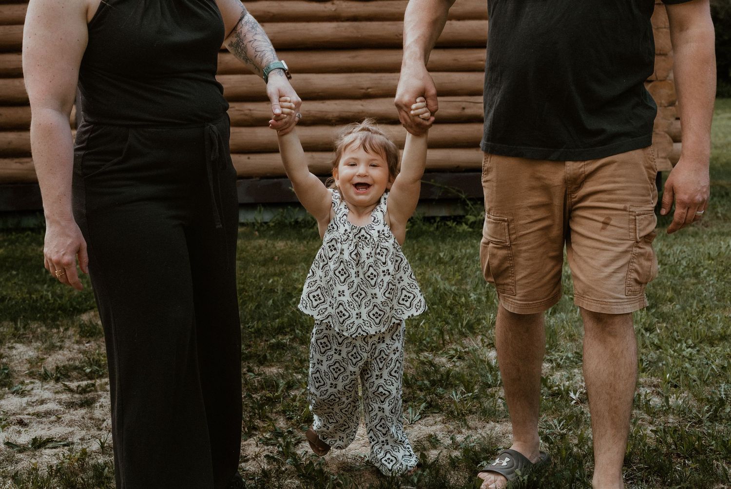 Happy toddler held by hands between parents outdoors near log cabin during family photo session.