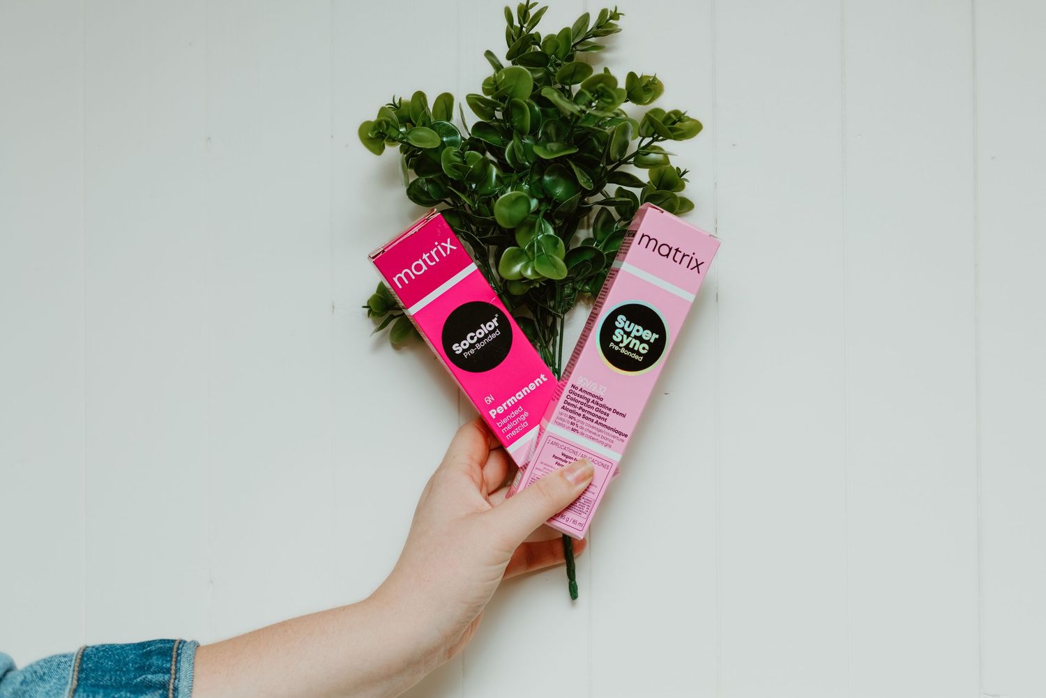 Hand holding two Matrix hair color tubes in pink packaging with a green plant on white background.