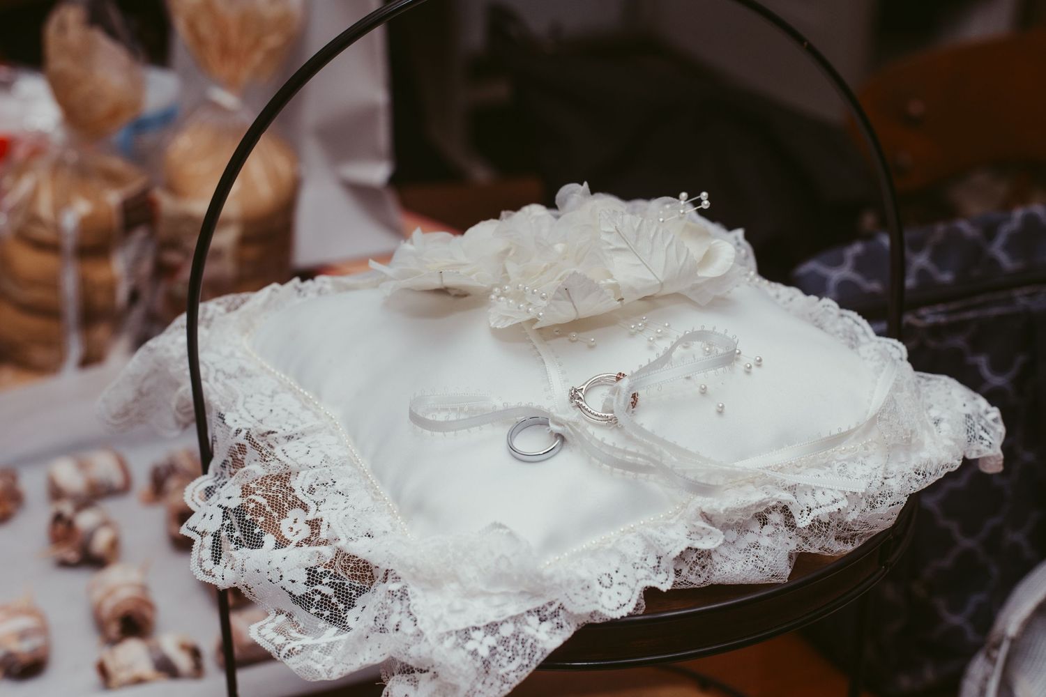 Wedding rings displayed on white lace ring bearer pillow at decorated reception dessert table.