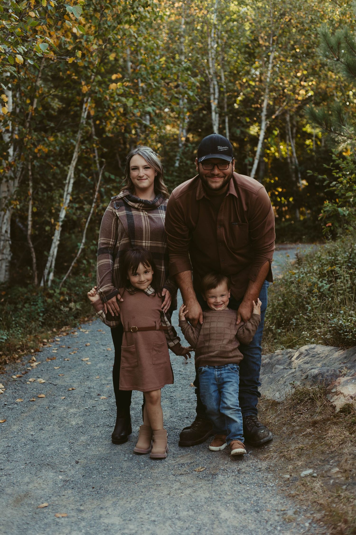 Family of four smiling together on a gravel path surrounded by colorful autumn foliage.