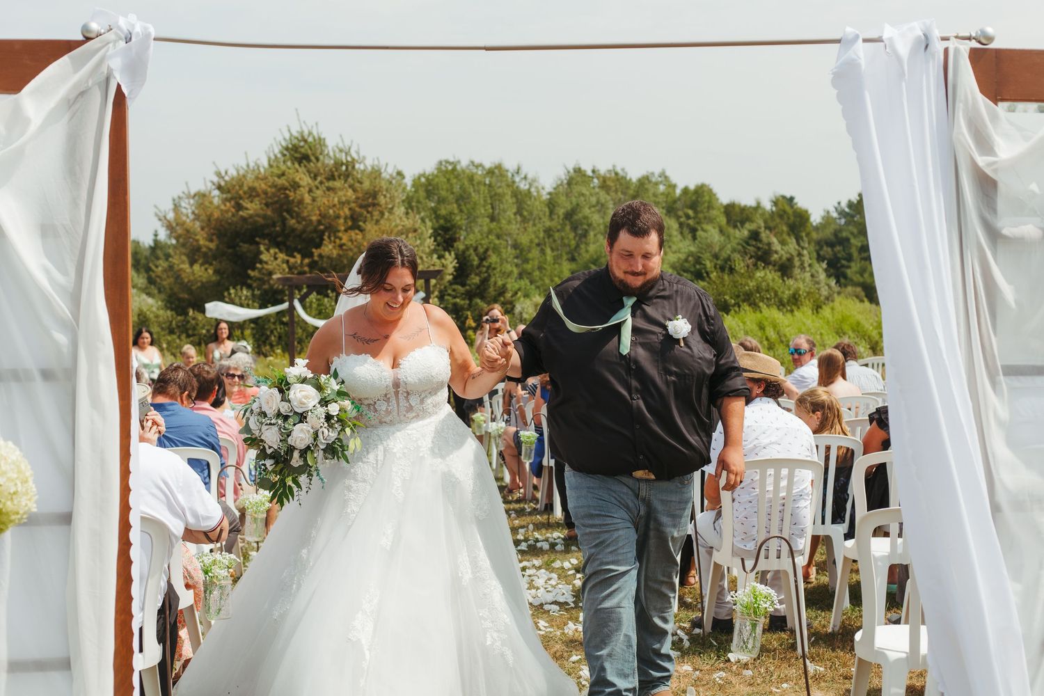 A bride and groom walk down the aisle together under a white draped wooden arch at an outdoor ceremony.