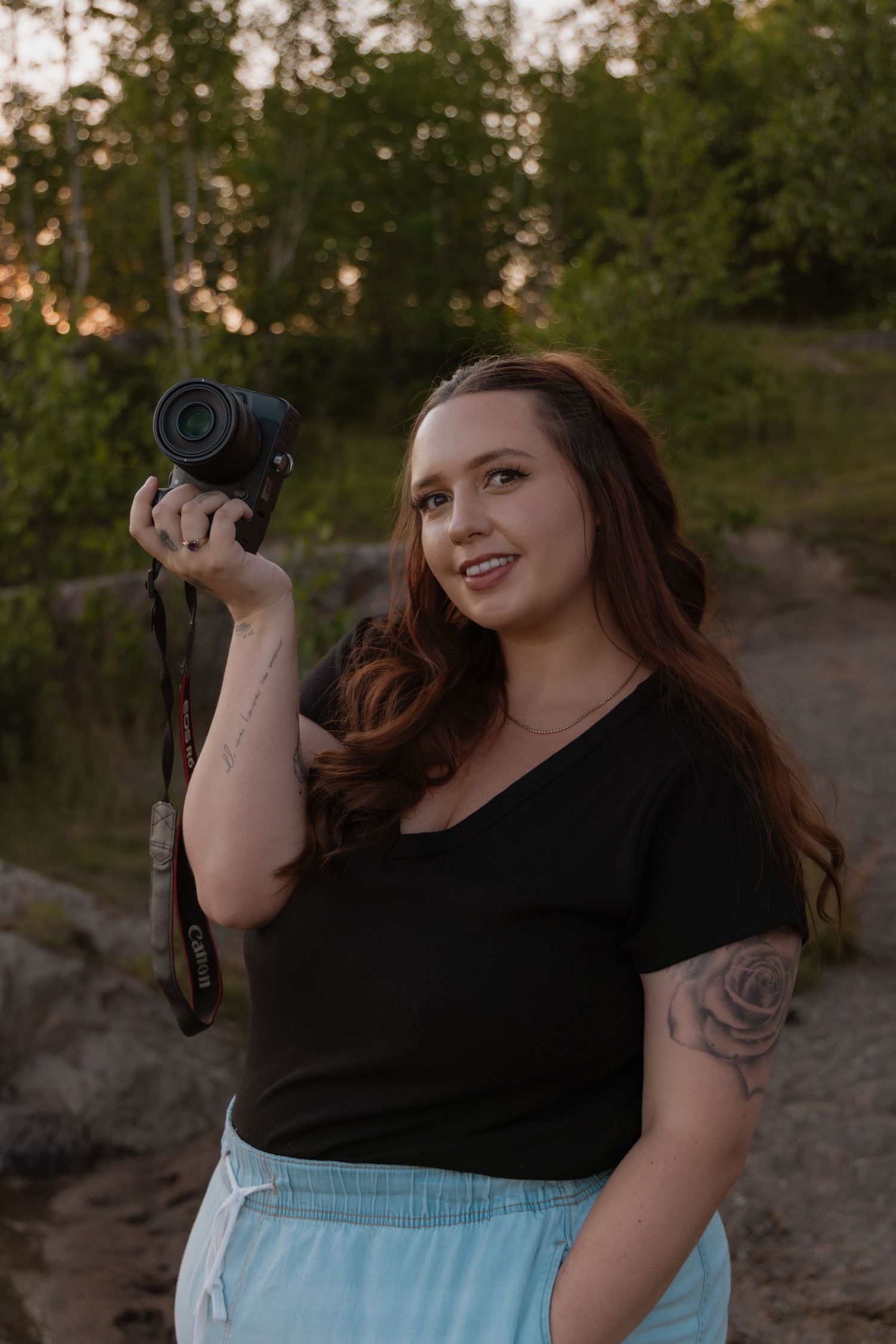 Woman with red hair holding a camera outdoors at sunset, wearing a black top and light blue pants with a rose tattoo.