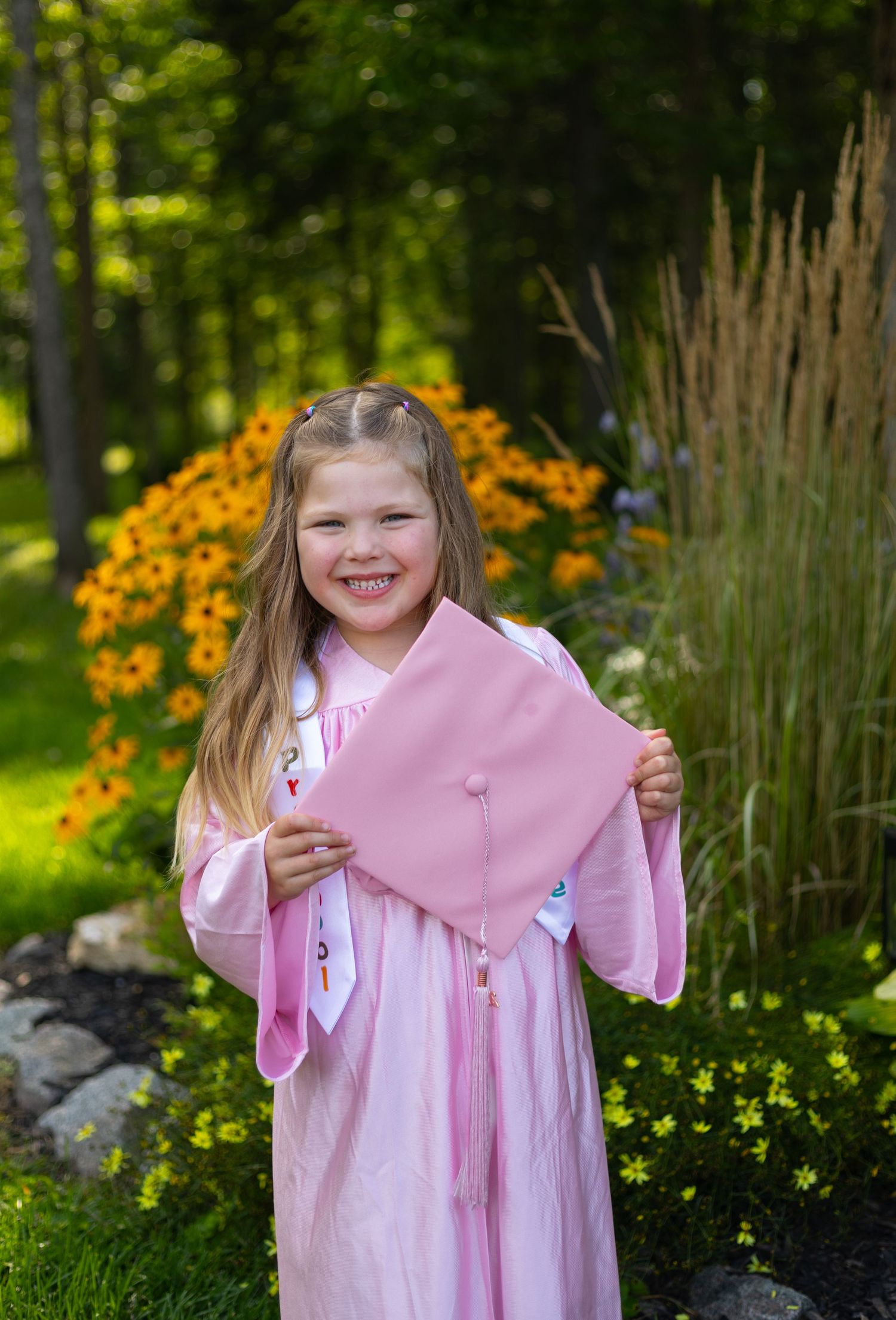 Young girl in pink graduation cap and gown smiling outdoors with colorful garden flowers in background.