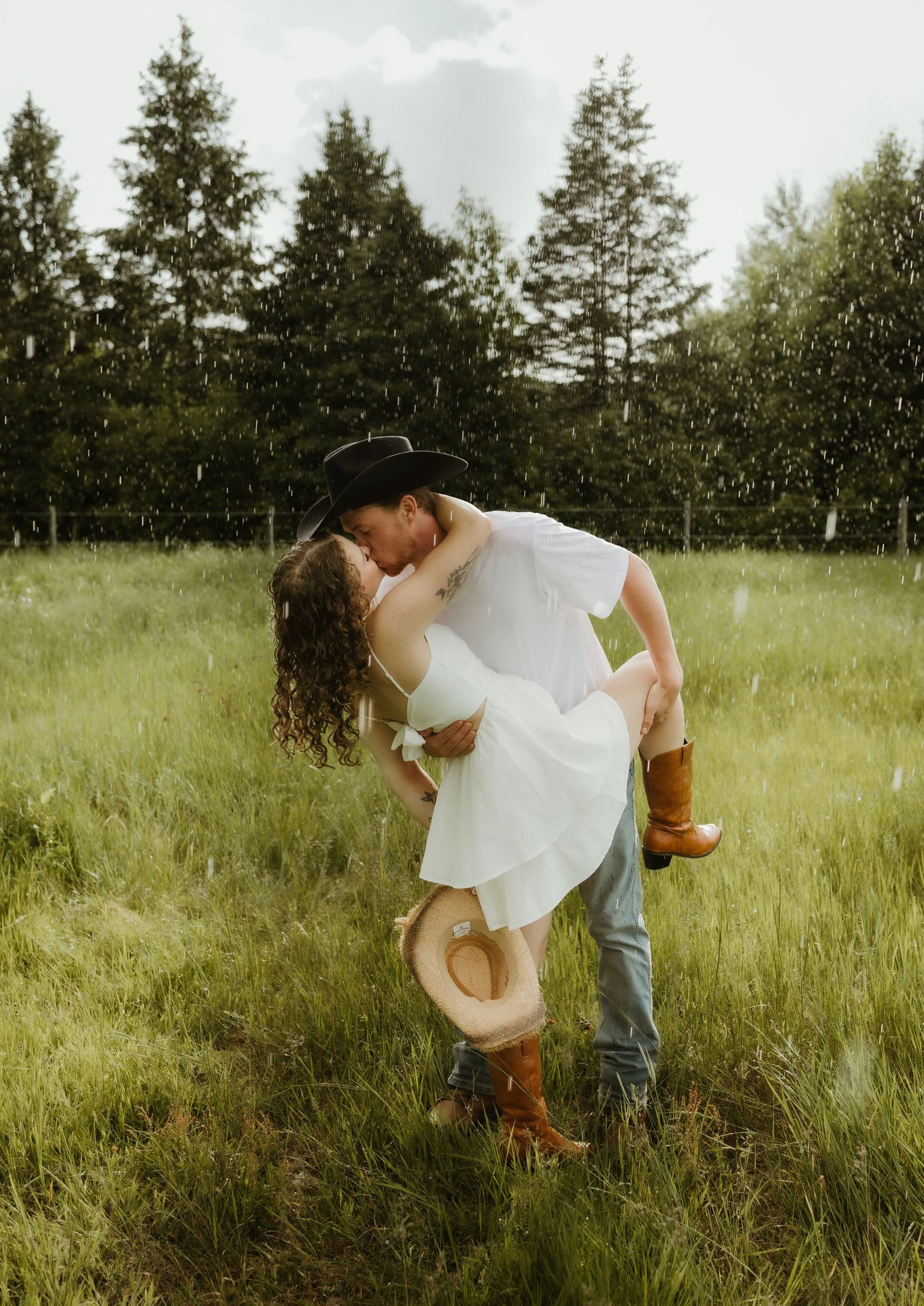 Couple sharing a romantic dip kiss in a rainy green field wearing cowboy hats and boots.