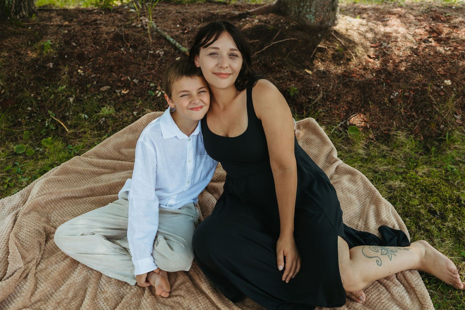 A woman in a black dress and young boy in white shirt sit together on a blanket outdoors, smiling warmly.