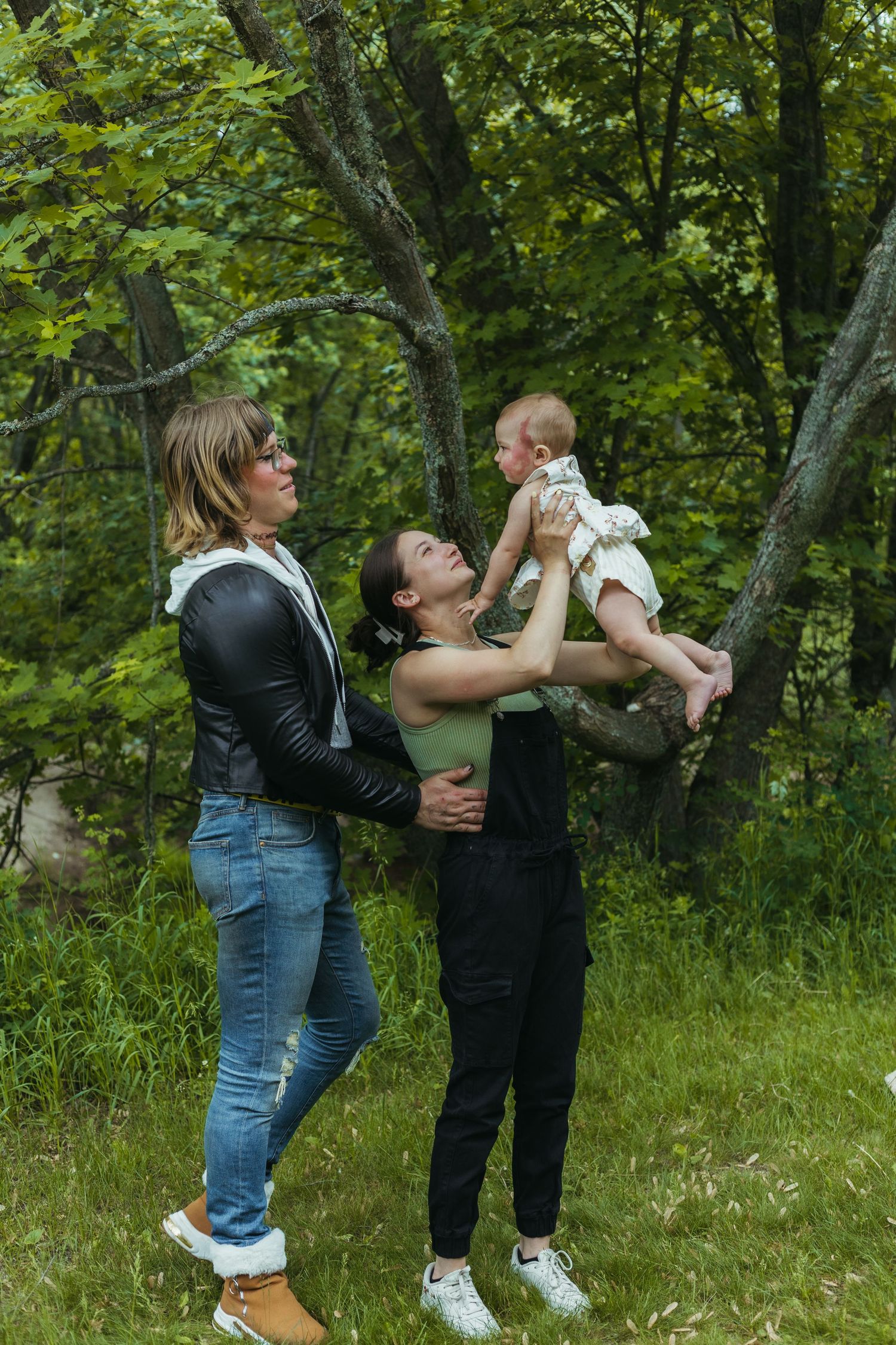 Two women playing with a baby outdoors in a lush green forest setting, one lifting the infant joyfully.