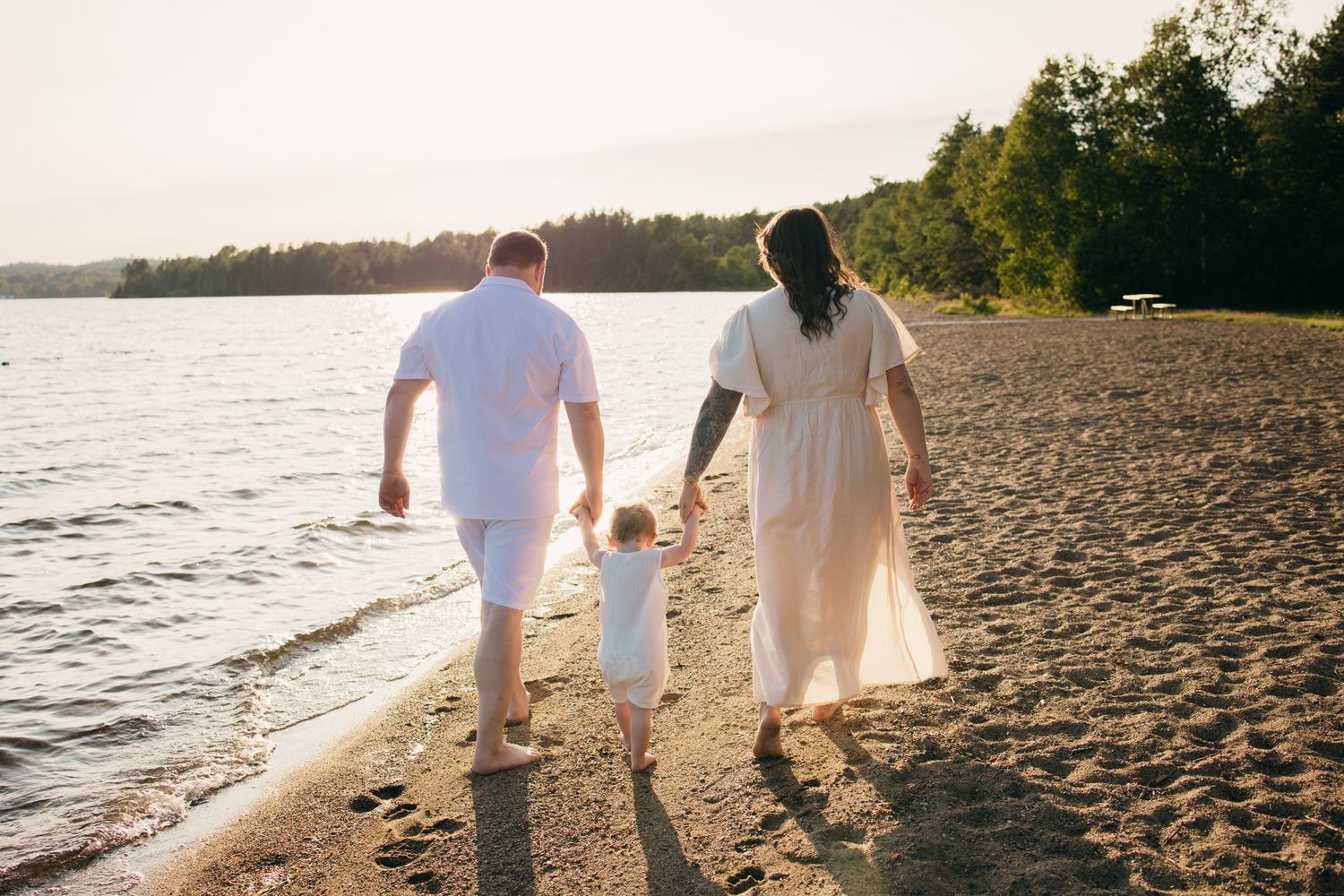 Family of three walking hand-in-hand along a sandy beach at sunset, with calm water and trees in the background.