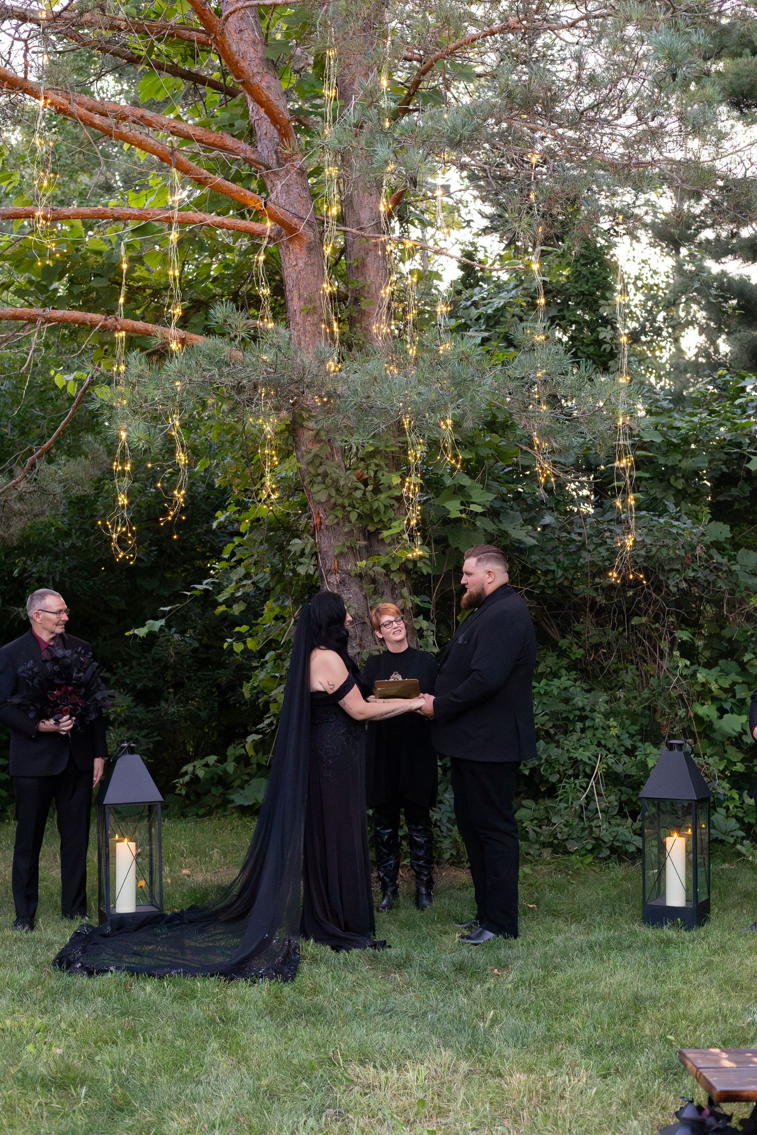 Gothic wedding ceremony under tree with hanging lights, black lanterns, bride and groom exchanging vows outdoors.